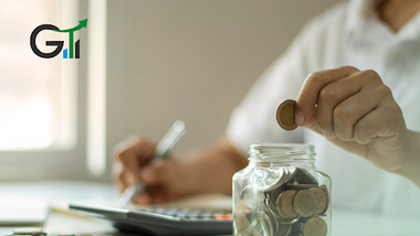 persona poniendo monedas en un frasco lleno de monedas y escribiéndolas en papel con calculadora en la mesa