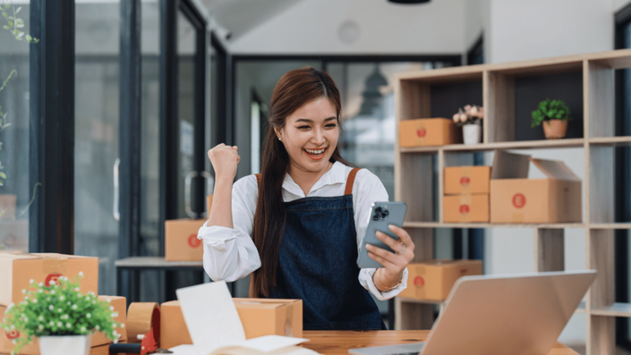 Emprendedora emocionada revisando su celular mientras maneja su negocio rentable en México de ventas en línea.